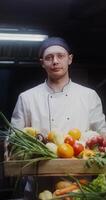 A man in a chef's uniform smiles looking at the camera while standing with a box of vegetables in his hands - looking photo