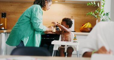 Mom feeds the baby sitting in a chair with yogurt, the eldest daughter uses a tablet sitting at the table in the kitchen - daughter photo
