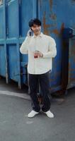 A young man of Chinese appearance is talking on a mobile phone, and drinking juice from a disposable cup with a straw standing near a metal wall outdoors photo