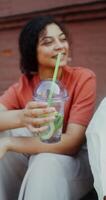 A woman drinks juice from a disposable cup with a straw while sitting on a city street, and talks to a person sitting next to her - sitting photo