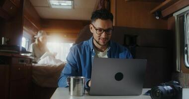 A young man uses a laptop and drinks a hot drink from a metal mug while sitting at a table in a van. A woman reads a book in the background - background photo