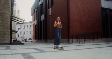 A young red-haired woman drinks lemonade while riding a longboard on a city street on a summer day - riding photo
