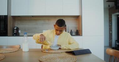 The boy has breakfast at home, he pours cereal into a plate while sitting at the table and looking at the tablet photo