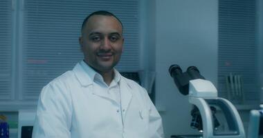 A man in a lab coat looks at the camera sitting in front of a microscope in a medical laboratory - microscope photo