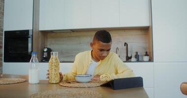 The boy has breakfast at home, he pours cereal into a plate while sitting at the table and looking at the tablet - sitting photo