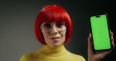 A woman with glasses and an orange wig smiles at the camera, holding a mobile phone with green screen in her hand, standing on a gray background in the studio - smiles photo