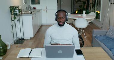 A man with headphones is talking on a chat on a laptop, sitting at a table at home. He writes in a notebook. Remote work photo
