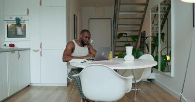 A man in pajamas uses a laptop and writes in a notebook sitting at a table in the kitchen at home with a serious look photo