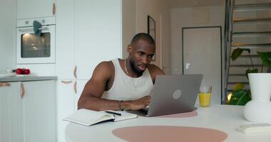 A man in pajamas uses a laptop and writes in a notebook sitting at a table in the kitchen at home with a serious look - writes photo