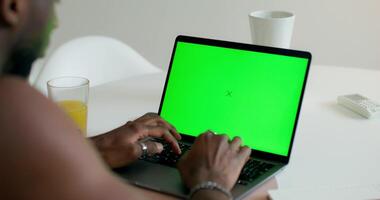 A man uses a laptop with a green screen sitting at a table at home, close-up of his hands, an unrecognizable person. Daylight photo