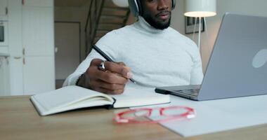 A man with headphones is talking on a chat on a laptop, sitting at a table at home. He writes in a notebook. Remote work - writes photo