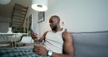 A man in pajamas uses a mobile phone and drinks water from a glass sitting on the couch at home. Daylight - drinks photo