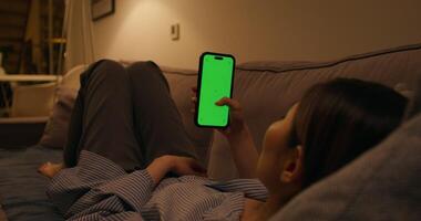 A woman uses a mobile phone with a green screen lying on the couch at home, an unrecognizable person photo