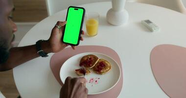 A man eats pancakes and uses a mobile phone with a green screen sitting at a table at home. Daylight - screen photo