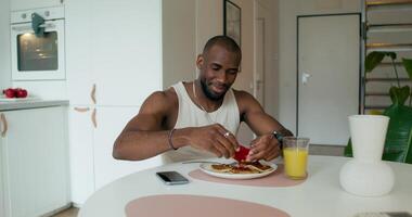 A man in pajamas squeezes jam out of a package onto pancakes while having breakfast, sitting at the table at home. Daylight - package photo