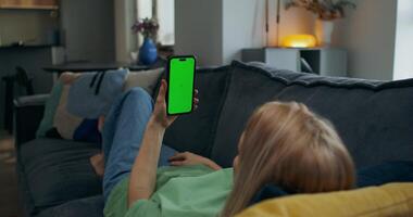 A woman holds a mobile phone with green screen in her hand. She is lying on the couch at home, rear view, unrecognizable person photo