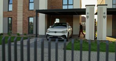 A man in casual clothes connects a charger to an electric car parked in front of a stylish cottage. A sunny day - connects photo