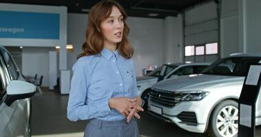 A car dealership manager in formal clothes smiles and talks to a customer standing near cars in a car dealership photo