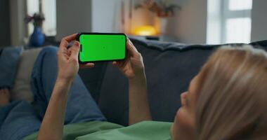 A woman holds a mobile phone with green screen in her hand. She is lying on the couch at home, unrecognizable person - unrecognizable photo