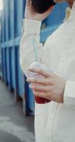 A young man of Chinese appearance is talking on a mobile phone, and drinking juice from a disposable cup with a straw standing near a metal wall outdoors - mobile photo