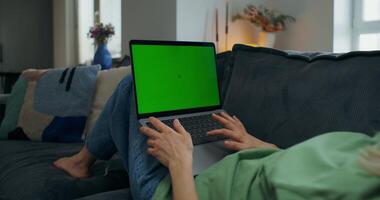 A woman uses a laptop with green screen lying on the couch at home, close-up of her hands, an unrecognizable person - person photo