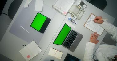 A laptop and a tablet with green screens are on the desktop in the medical office. The doctor fills out medical documents, only his hands are in the frame, unrecognizable photo