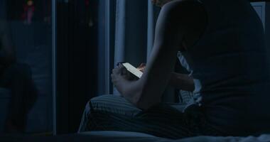 A man uses a mobile phone while sitting on the bed at night. Close-up of his hands, an unrecognizable person. Dark room, home interior - unrecognizable photo