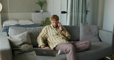 A bearded man uses a laptop and talks on a mobile phone sitting on the couch at home with a serious look. Daylight photo