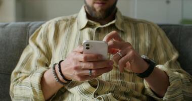 A man is typing on a mobile phone sitting on the couch at home. close-up of his hands, an unrecognizable person. Daylight - sitting photo