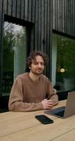 A man in casual clothes smiles looking at the camera sitting in front of an open laptop on the terrace of a stylish cottage on a summer day - smiles photo
