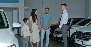 The car dealership manager shows cars to customers standing in the showroom. A family with a child chooses a car - standing photo