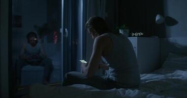 A man in pajama pants uses a mobile phone while sitting on the bed at night. Dark room, home interior photo