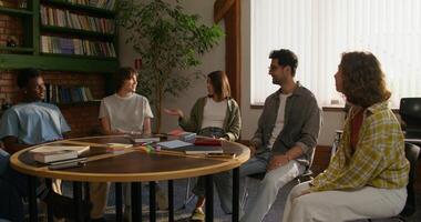 A group of young people are smiling and talking while sitting around a round table indoors. Notebooks and books on the table. Daylight - talking photo