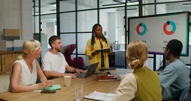 A group of young people are sitting at a table in an office meeting. A female speaker is talking standing near the screen. An open laptop is in front of a man. Daylight photo