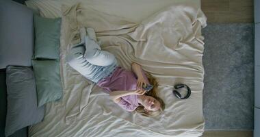 A young woman in casual clothes uses a mobile phone lying on a bed at home. Top view, daylight - mobile photo