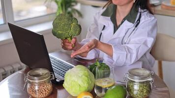 A doctor conducts an online consultation on nutrition at a table with fruits and vegetables. Eating broccoli helps maintain health. The concept of health, medical practice, fitness. video
