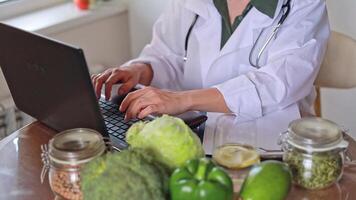 A nutritionist doctor works at a table with vegetables and fruits, types text on a laptop keyboard and communicates with patients, makes a meal plan and diet. The concept of health, clinic, wellness. video