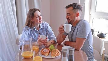 A couple enjoys a romantic breakfast with croissants and coffee, having a cheerful conversation at home. video