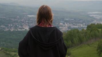 Young woman in black hoodie standing on hilltop, overlooking expansive valley with distant town amid verdant forest, embodying peaceful exploration and personal achievement video