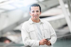 A young man stands confidently with a smartphone in hand, engaging with advanced facial recognition technology in a contemporary urban setting. This reflects the new normal of digital identification. photo