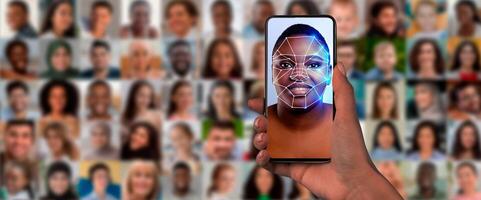 A hand holds a smartphone displaying a digital face scan. Behind, a collage of diverse faces highlights the technology's capability for identification in modern society. photo