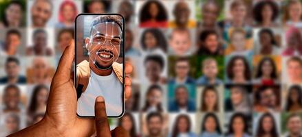 A hand holds a smartphone displaying a smiling person face. Behind, a collage of diverse portraits highlights the advanced facial recognition technology used for identification in various settings. photo