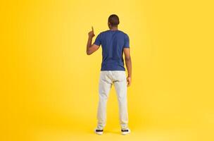Back View Of African American Man Pointing Finger Up Pushing Invisible Button Or Touchscreen Standing Over Yellow Studio Background. Look Upward. Full Length Shot photo
