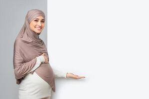 Pregnant muslim woman in hijab pointing at white advertisement board with free space, touching her belly and looking at camera, grey background photo