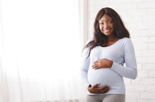 Beautiful happy pregnant black woman touching her tummy, staying next to window, posing at home, free space photo
