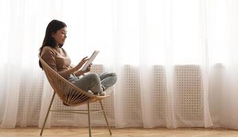 Lazy weekend. Girl using tablet, sitting in wicker chair against window, panorama with empty space photo