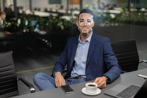 A man dressed in business attire sits comfortably at a conference table, using facial recognition technology for identification while enjoying a cup of coffee in a contemporary office environment. photo