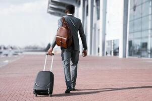 Business Trip Concept. Black Businessman With Travel Suitcase And Backpack Walking Back To Camera Leaving Airport. Copy Space photo