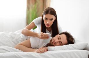 Infidelity. Shocked Wife Reading Message From Lover On Phone While Cheating Husband Sleeping In Bedroom At Home. Jealous Woman Checking Boyfriend's Cellphone Chats. Affair, Relationship Issues photo