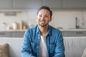 A man smiles while sitting on a couch in a contemporary kitchen, where facial recognition technology is being used to verify his identity. The modern design reflects a new normal in tech. photo